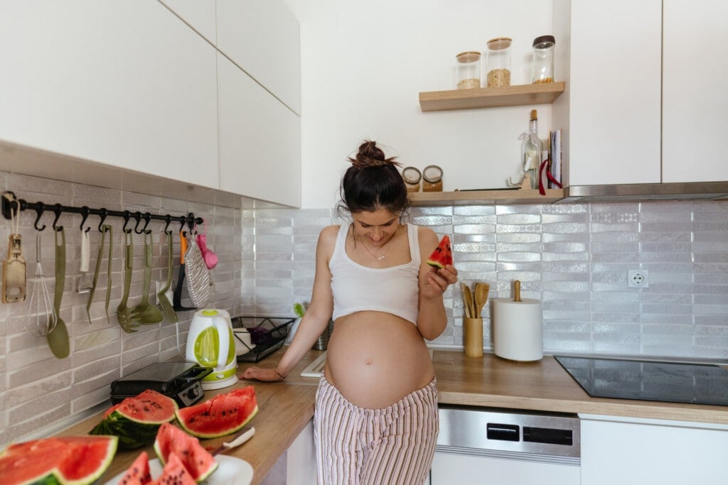 Photo of a smiling pregnant woman eating watermelon in the kitchen of her apartment; enjoying and craving food during pregnancy.