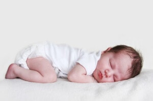 Newborn baby sleeping on his tummy wearing a white onesie.