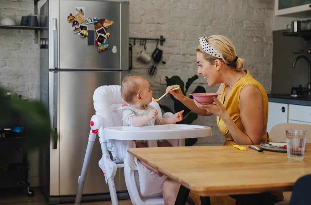 An adorable Caucasian baby being fed solid food by her beautiful young smiling Caucasian mother in a kitchen at home.