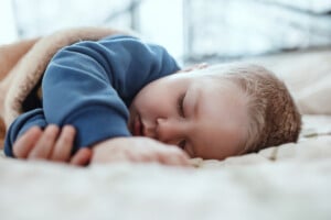 A young child with short hair sleeps on a bed, covered with a beige blanket, wearing a blue long-sleeve top.