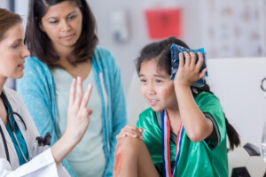 Serious female ER doctor holds up three fingers while talking with injured female elementary age soccer player. The doctor is asking the girl how many fingers she is holding up. The girl is wearing a medal and soccer uniform.