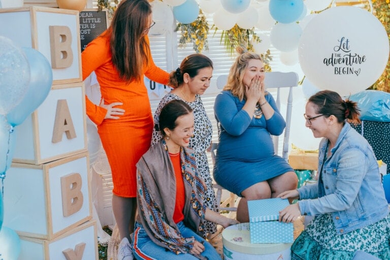 Female friends open gift boxes with a pregnant woman at a baby shower. Party decorations in white and blue colors, baby boy