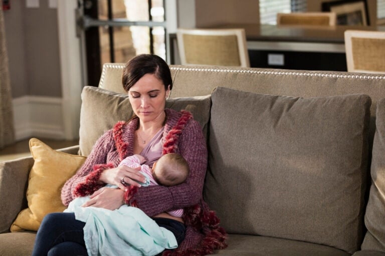 A mid adult woman in her 30s, holding her beautiful 2 month old baby girl, sitting on a sofa at home, nursing. The mother has a serious expression on her face, eyes closed. Perhaps she is tired, or a little sad.