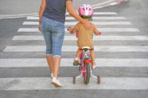 An adult and a young child are crossing a street on a pedestrian crossing. The adult, wearing a blue shirt and denim shorts, holds the child, who is wearing a pink helmet and riding a bicycle with training wheels. Following safety rules, the child's bike is pink and red for visibility.