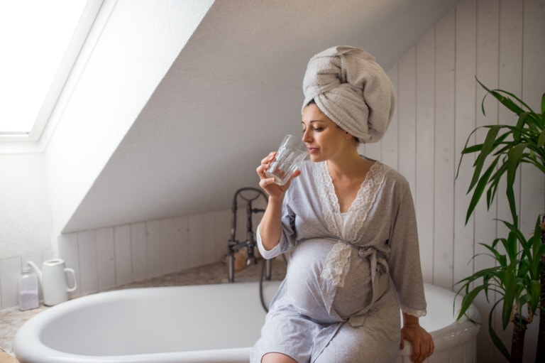 Portrait of happy pregnant woman indoors in bathroom at home, drinking water.