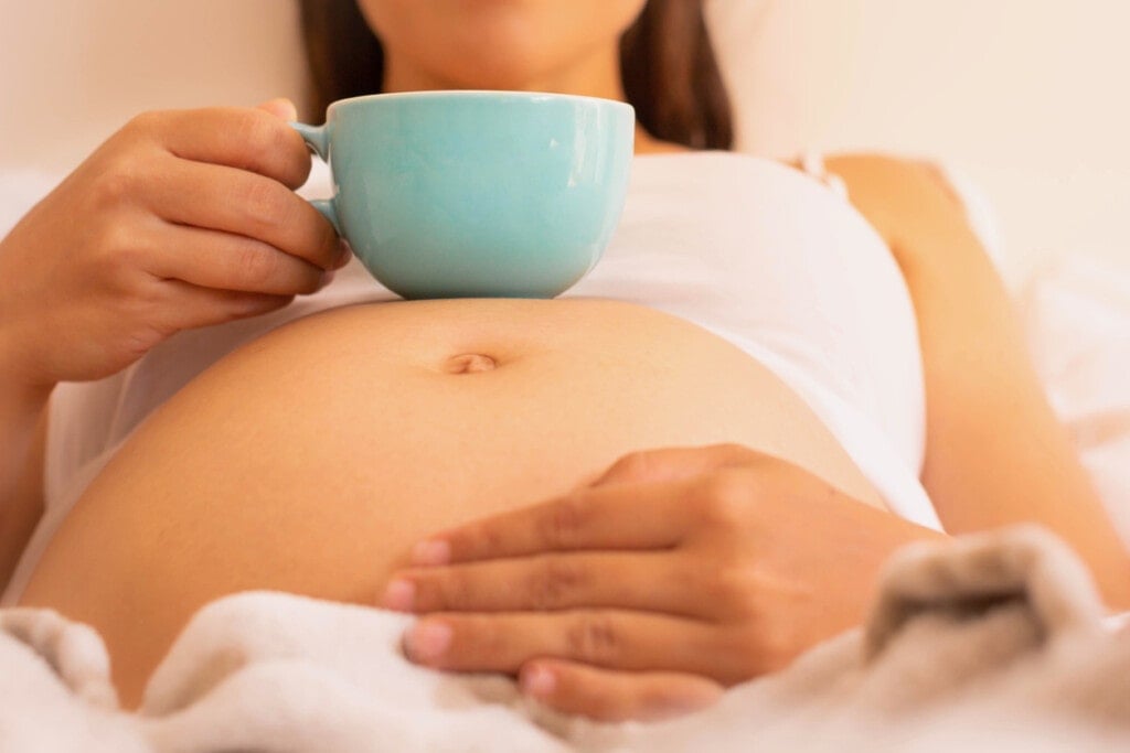 A close up of a pregnant baby belly, woman holding a mug with coffee or tea, and laying in bed.