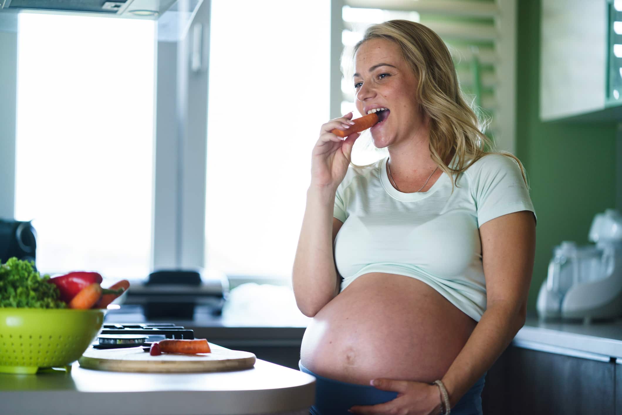 Young pregnant woman eating a carrot in the kitchen and looking away