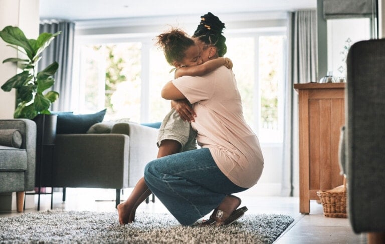 An adult kneels and hugs a child in a modern living room with large windows allowing natural light. The room, decorated with a plant, armchairs, and a wooden cabinet, sets the scene as they share a warm embrace. It seems like an apology being made after family arguments; both appear relieved and connected.