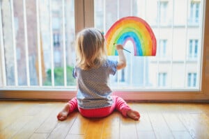 A busy toddler with blond hair, wearing a blue polka-dotted shirt and red leggings, sits on the floor facing a large window. The child is holding up a drawing of a rainbow against the window. The background shows buildings outside.