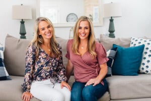 Two women sitting on a beige sofa in a cozy living room, smiling and sharing tips on how to get your baby talking. Both have long blonde hair. The woman on the left wears a patterned top and white pants; the woman on the right wears a maroon top and blue jeans. The background includes cushions and lamps.