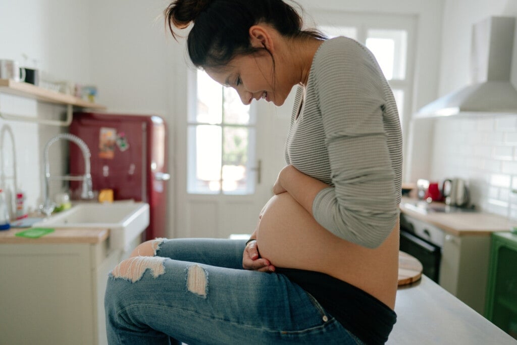 Photo of a smiling expectant mother looking at her baby bump and enjoying her morning in the kitchen of her apartment; the daily routine of a pregnant woman.