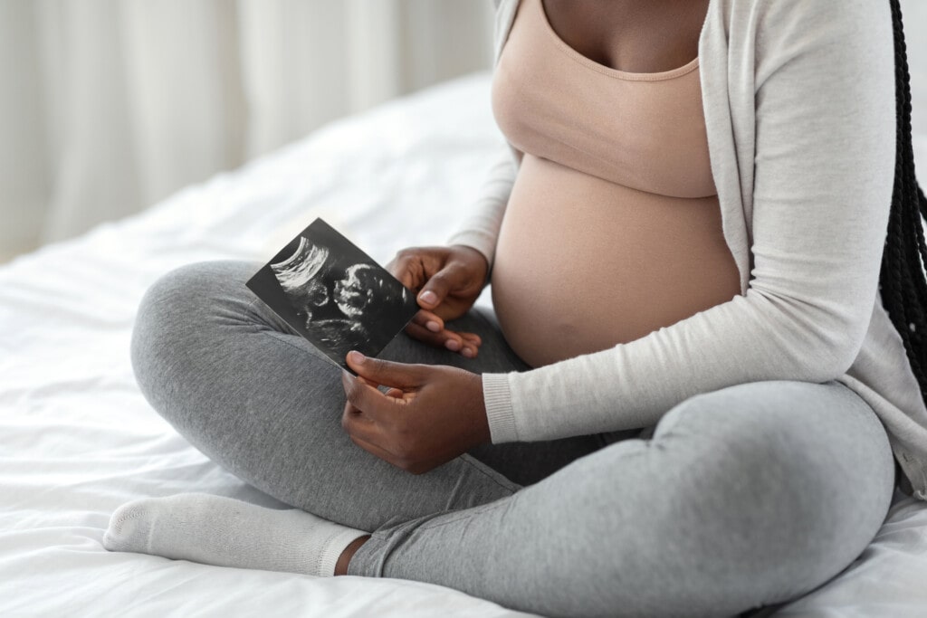 Pregnant african american woman looking at ultrasound scan of baby at home, holding sonography image in hands, sitting on bed, enjoying healthy pregnancy, cropped image, closeup
