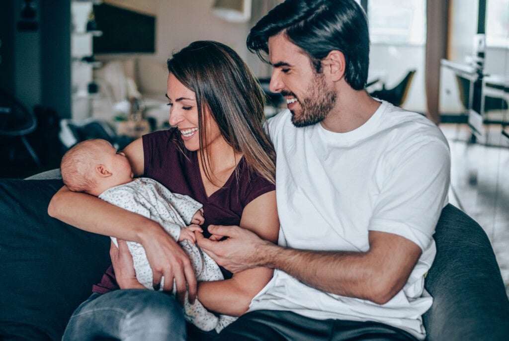 Beautiful young couple embracing their newborn baby on the sofa. Smiling young parents with their baby girl at home.