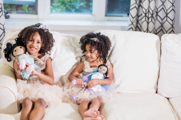 Two cute mixed race African-American and Hispanic girls, sisters 3 and 5 years old, playing together, sitting side by side on a sofa, holding dolls in their laps, wearing princess dresses and tiaras, smiling at the camera.