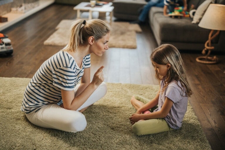 A woman with blonde hair, wearing a striped shirt and white pants, is sitting on a green rug, holding up a finger while talking to a young girl with brown hair. The girl, sitting cross-legged and looking down in the living room, seems to be listening intently as if awaiting the last word.