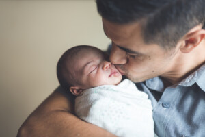 An ethnic dad gently kisses his newborn daughter on the cheek. He is holding her cradled in his arms as she is swaddled up comfortably for her nap.