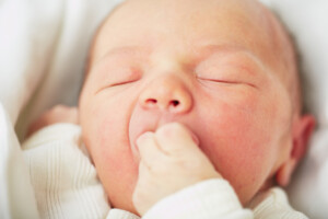 Newborn baby is peacefully sleeping on white blanket with his hand in his mouth.