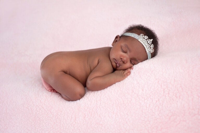 Portrait of a one month old, newborn, baby girl. She is wearing a rhinestone crown headband and sleeping on a soft, pink blanket.