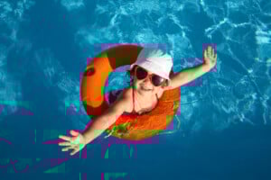 A child is floating in a swimming pool using an orange inflatable ring. The child is wearing a white hat, red sunglasses, and a red swimsuit, with arms outstretched while smiling up at the camera. This snapshot perfectly captures one of the fun summer activities for kids in clear, blue water.