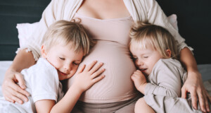 Pregnant woman with her children relaxing in bed. Loving mother and toddlers together at home. Little kids hold hands on pregnant belly of mom.