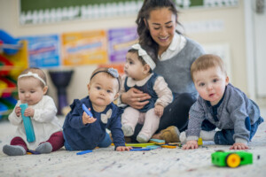 A group of babies are indoors in a day care centre. They are sitting on the carpet and playing with toys.