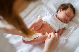A newborn baby lies on a white cloth as an adult changes their diaper. The babys umbilical cord stump is visible.