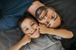 Dad and son lying down together smiling, with the son playfully stretching his mouth into a goofy grin