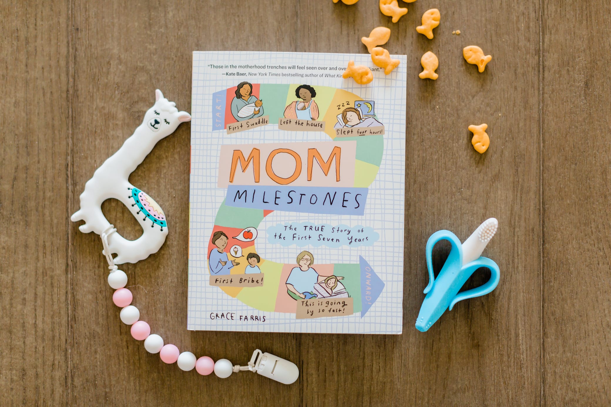 Mom milestones book on a table with some goldfish crackers, a teether, and pacifier clip laying next to it.