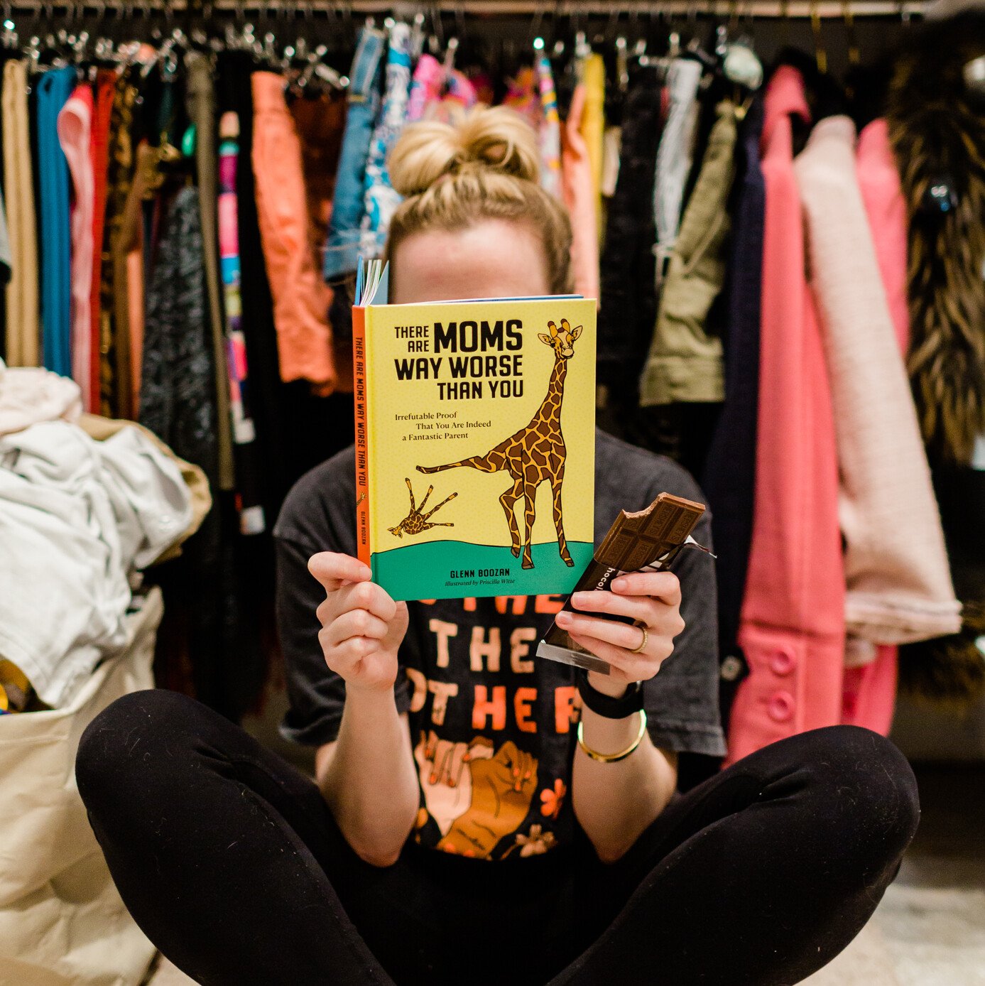 Woman sitting on the floor in her closet reading a book and holding a chocolate bar.