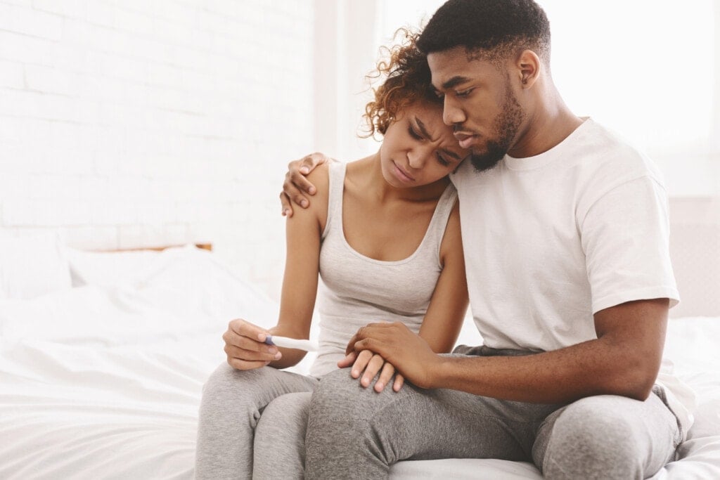 Vấn đề vô sinh. Cặp vợ chồng người Mỹ gốc Phi thất vọng ngồi trên giường với kết quả thử thai âm tính.