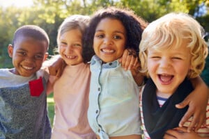 Four children stand outdoors with their arms around each other, smiling broadly at the camera. They are in a park or garden setting with green trees in the background, embodying perfect summer playdate ideas. The children are wearing casual clothes and appear to be enjoying a sunny day.