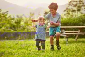 A young boy and a toddler holding hands while playing on a grassy field. The boy is smiling and wearing a gray t-shirt and blue shorts, while the toddler is dressed in a blue dress and gray leggings. A wooden picnic table is visible in the background, capturing how to raise kind kids in every moment.