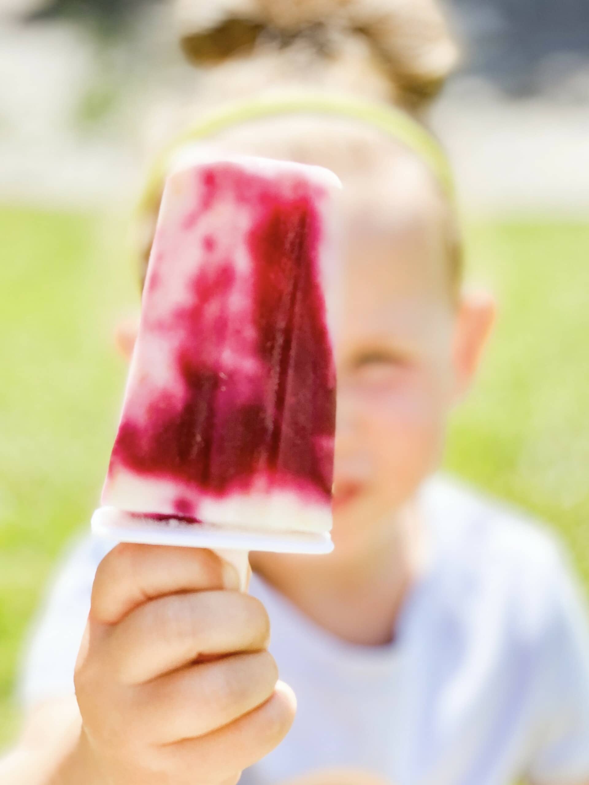 A child holding a homemade fruit and yogurt popsicle with vibrant red and white swirls, reflecting one of our best popsicle recipes. The blurred face in the background hints at joy, set against a grassy area on a sunny day.
