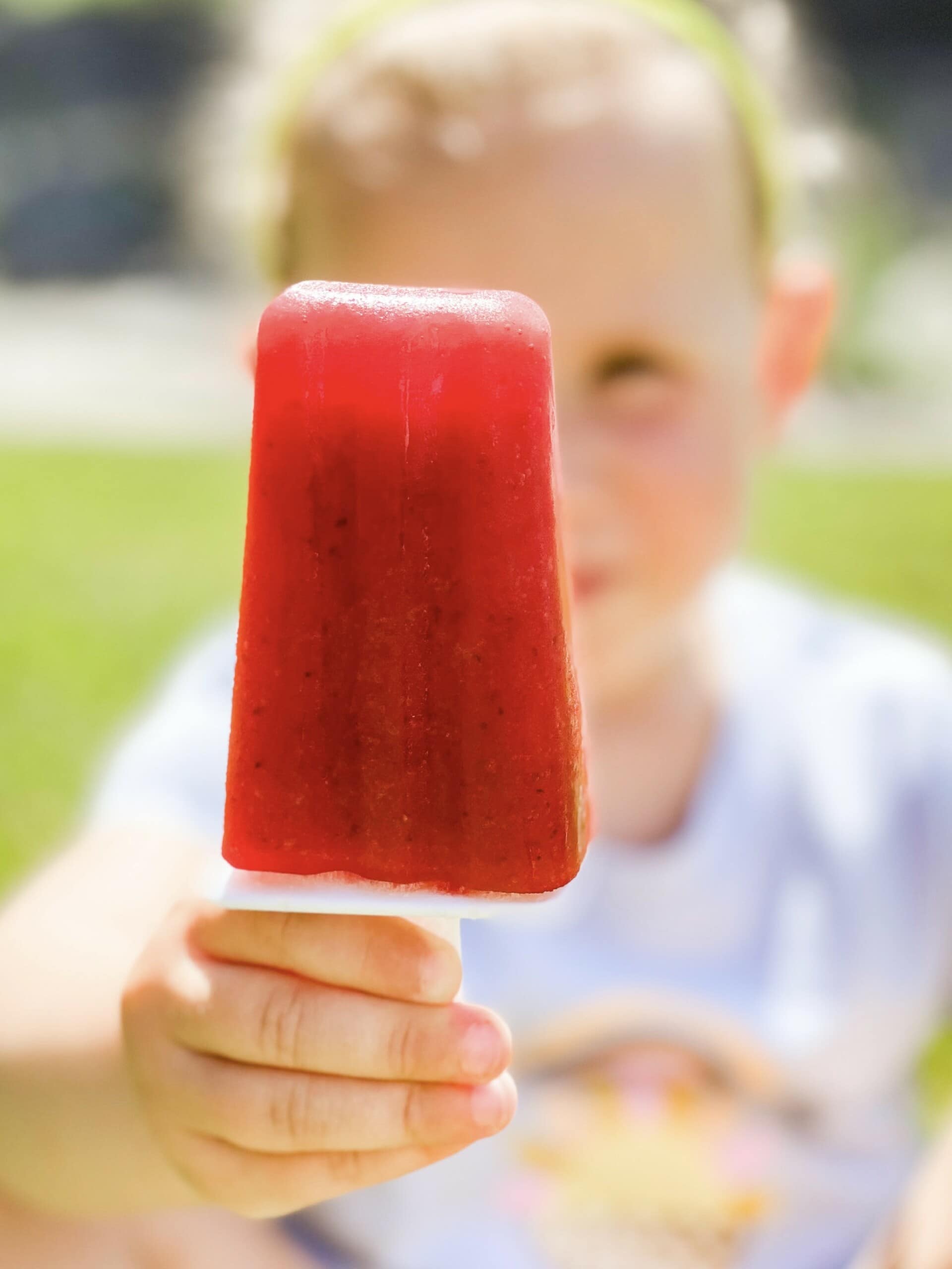 A child wearing a white shirt holds a red popsicle up to the camera with their right hand. The background is blurred, with green grass visible in some areas. The child's face is out of focus, but their delighted expression hints at the joy of summer and discovering new popsicle recipes.