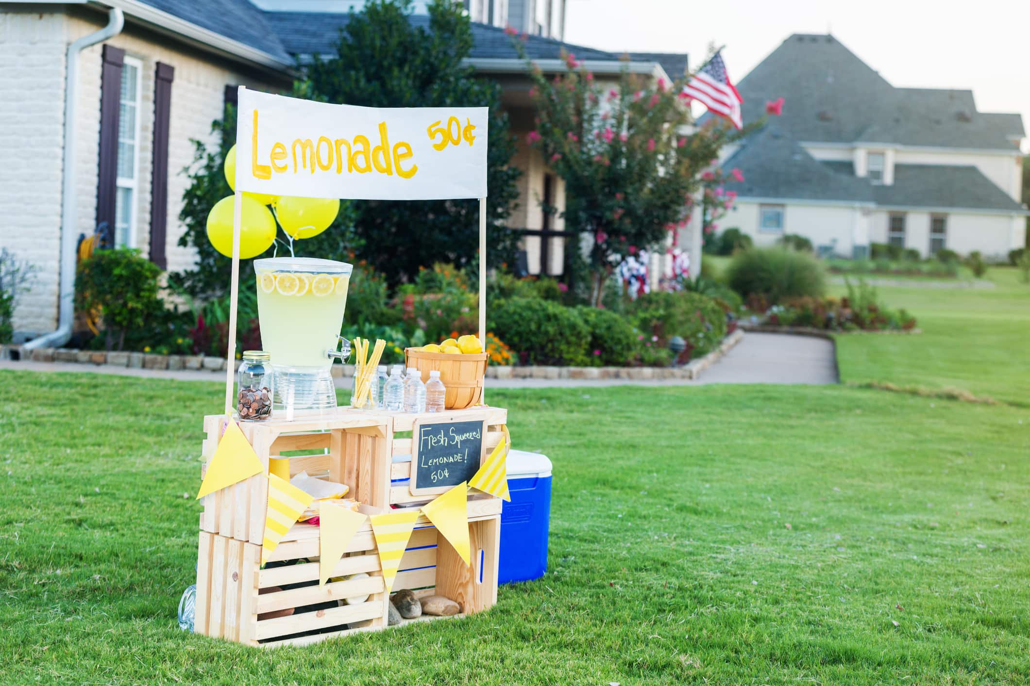 A refreshing lemonade stand is set up in the front yard of a home in the suburbs on a sunny summer day. A beverage dispenser, basket of fresh lemons and glasses are on the stand. Balloons are attached to the stand. A banner across the top of the stand lists the price of the lemonade.