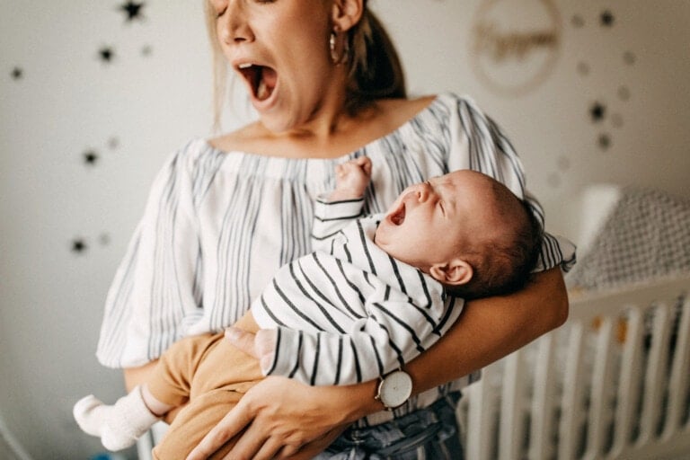 Mother holding her baby son. She is yawning while he is crying.