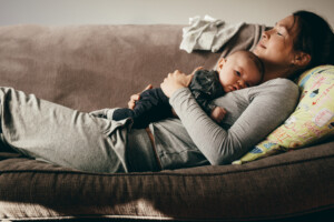 Side view of a woman lying on couch with eyes closed and holding her baby on her chest. Tired mother taking rest sleeping on a sofa with her baby on her.