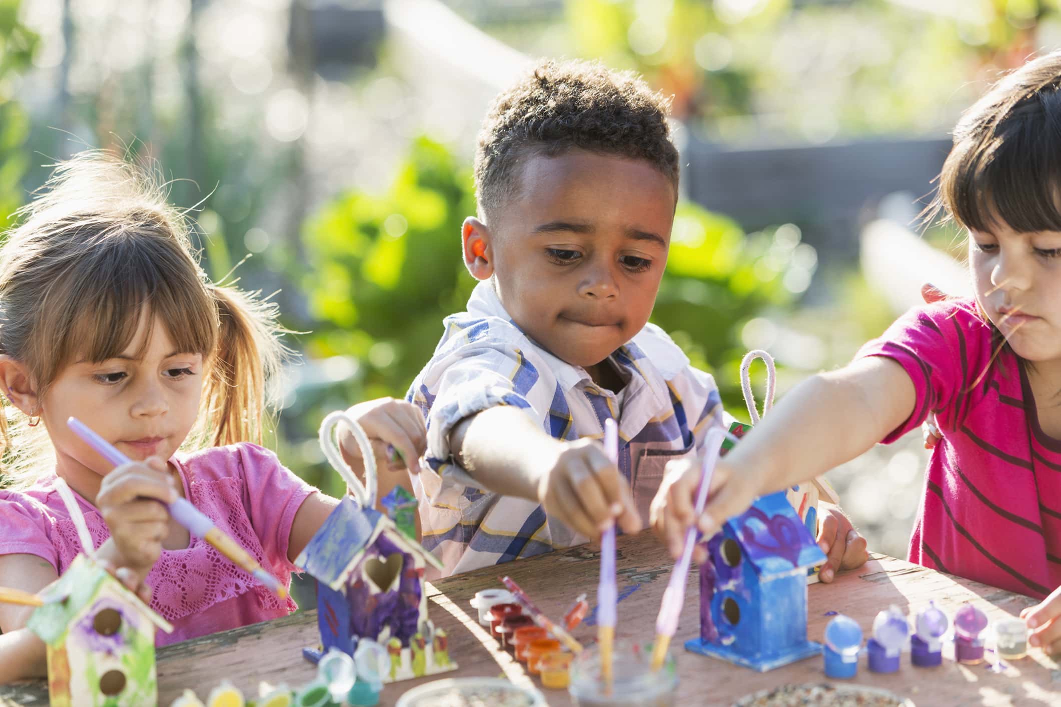 A multiracial group of three serious young children doing an arts and crafts project, painting little wooden bird houses for Earth Day. It is a bright, sunny day and they are sitting at a wooden table reaching with their paintbrushes.