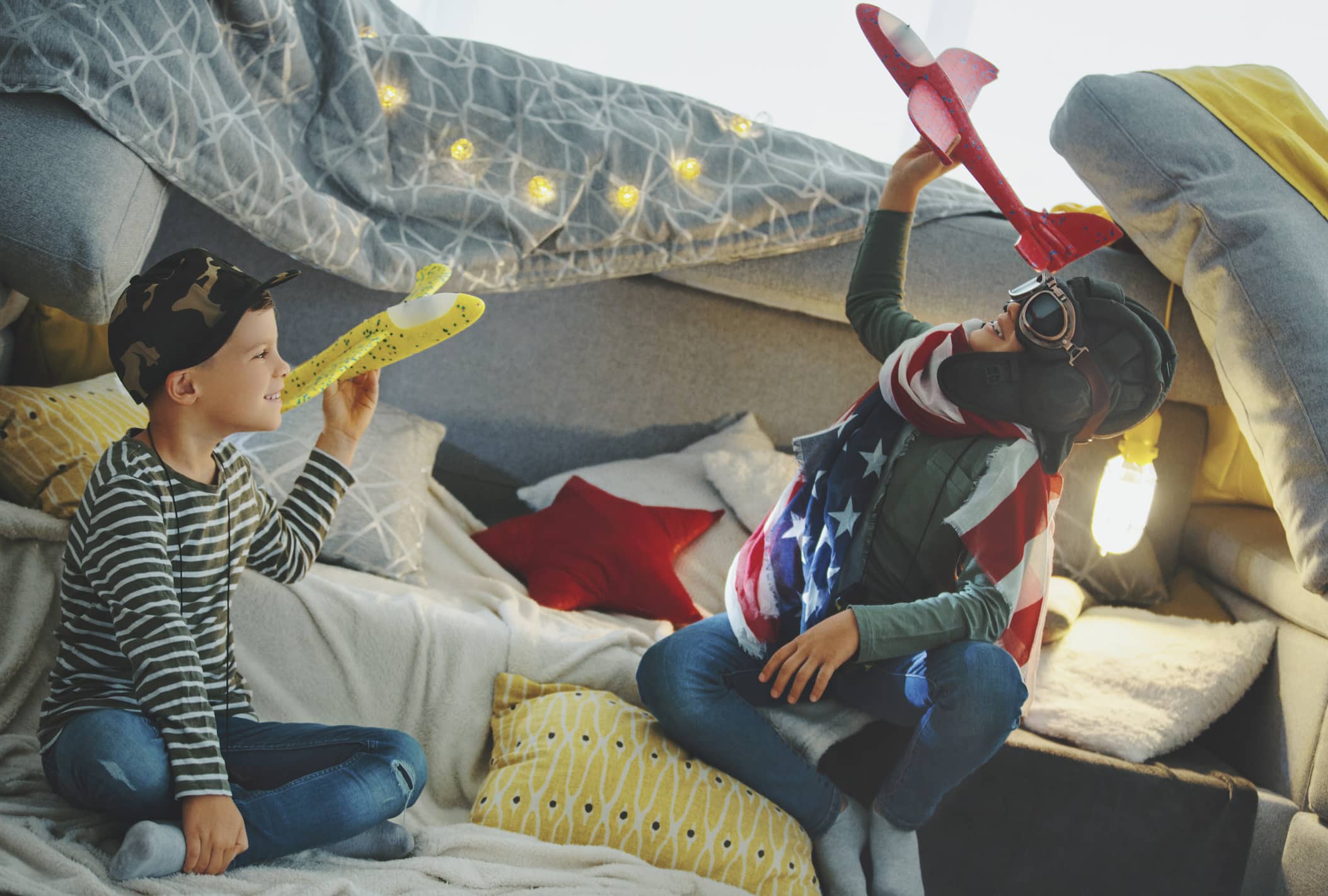 Brothers are playing in the living room. They were disguised as military pilots. They are sitting on a fortress they made of a sofa. And they fly with their toy planes.