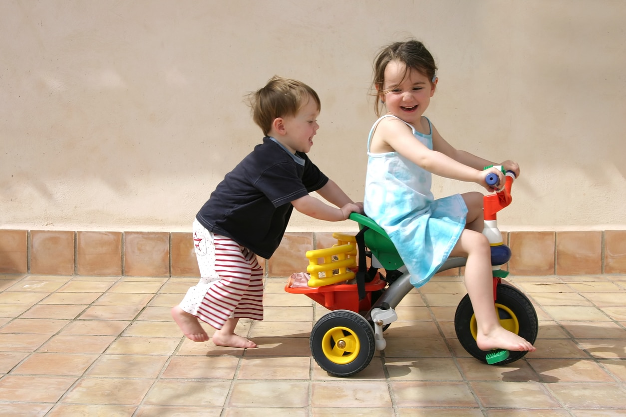 A small boy pushes a small girl on a tricycle