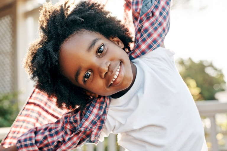 A young child with curly hair smiles and tilts their head, showcasing their vibrant personality. They are wearing a white shirt and have a plaid shirt draped over their shoulders. The background shows outdoor greenery and a wooden fence, with sunlight filtering through.
