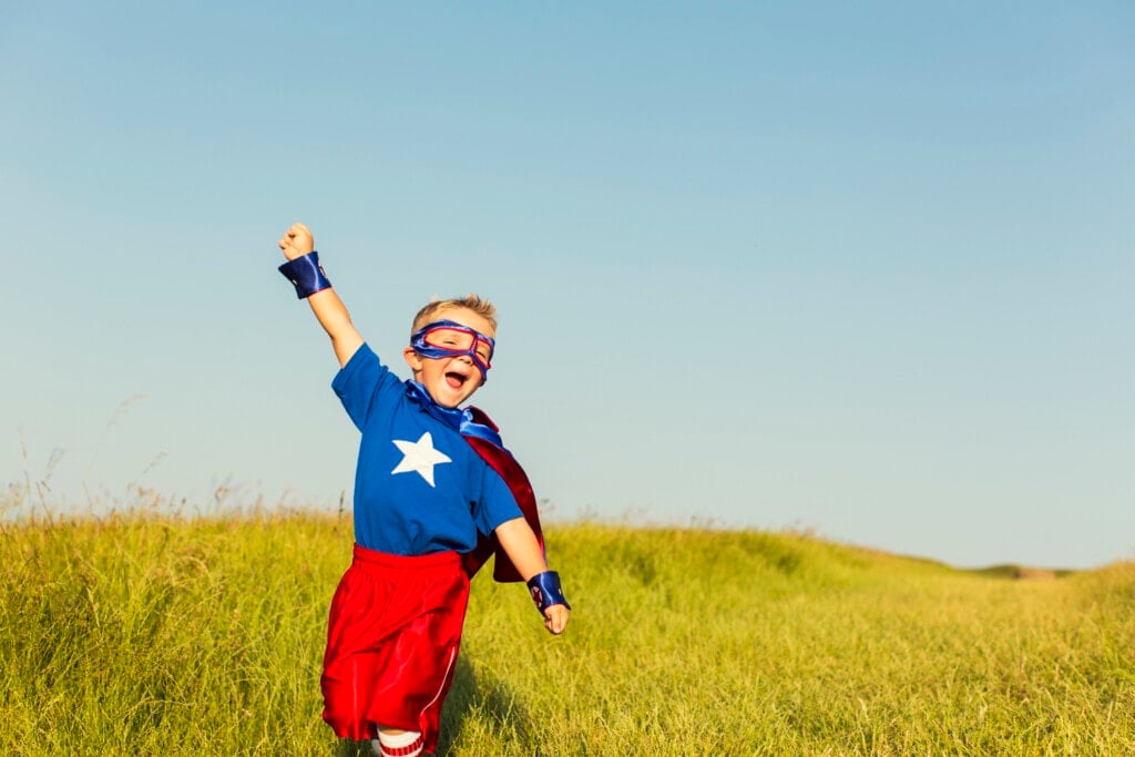 A young boy wearing a superhero cape and mask is ready to face life's challenges with confidence. He has his arm raised to the sky while standing in tall grass.