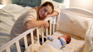 A woman in a beige robe is leaning on the side of a white crib, looking tired with eyes closed. Inside the crib, a baby dressed in a light blue outfit is lying on a striped mattress with a cream-colored blanket partially covering them. It's clear the baby wakes every hour from her weary expression.