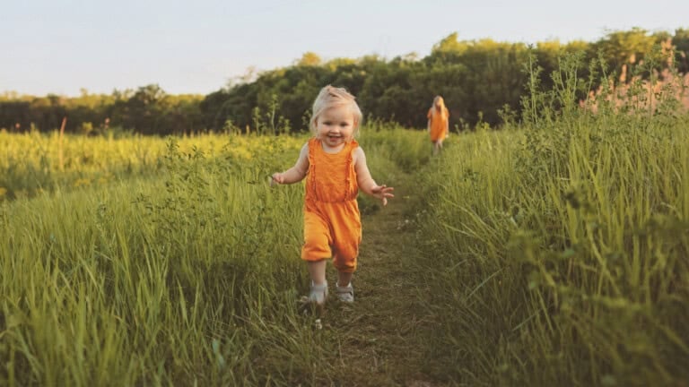 A young child wearing orange overalls runs down a grassy pathway in a green field. Another person in the distance, also clad in orange, strolls along the same path. The serene scene is set in a countryside area under a clear sky, evoking timeless country girl names.