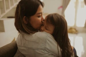 A woman sits on a couch, gently kissing a young child on the forehead while holding them in her arms.