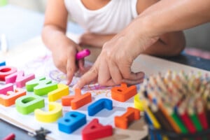 A child and an adult are engaging with colorful alphabet letters on a table. The child is holding a pink marker and drawing on paper. Nearby, there is a container filled with colored pencils. As the adult points at one of the letters, they smile, showing how fun it can be to teach your child the alphabet.