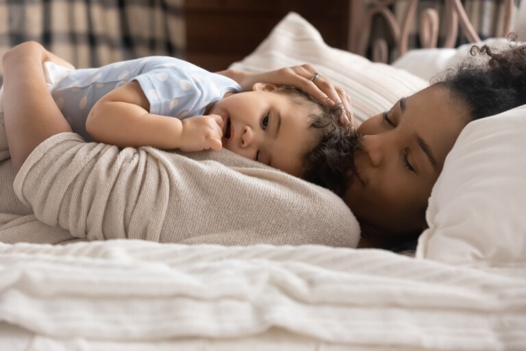 Happy loving young African American mom lying in bed caress cuddle little toddler daughter kid. Caring biracial mother relax in bedroom at home lull small baby girl child to sleep.