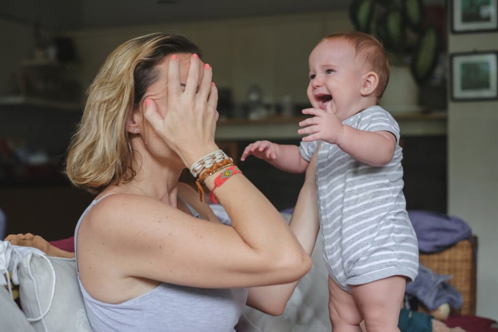 Shot of a frustrated Caucasian mother feeling postpartum depression.She's covering her face with her hand as she's coping with her cranky baby boy. She supports her crying baby to stand with one hand in front of her.