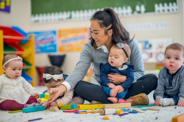 a young female teacher sits cross-legged on the floor while she plays with a few babies. She holds on in her lap while she passes some blocks to another. A little boy is off on the side drooling.