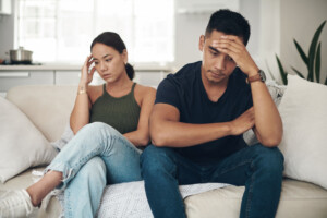 A man and a woman are sitting on the couch next to each other with their heads in their hands feeling overwhelmed by a difficult conversation.